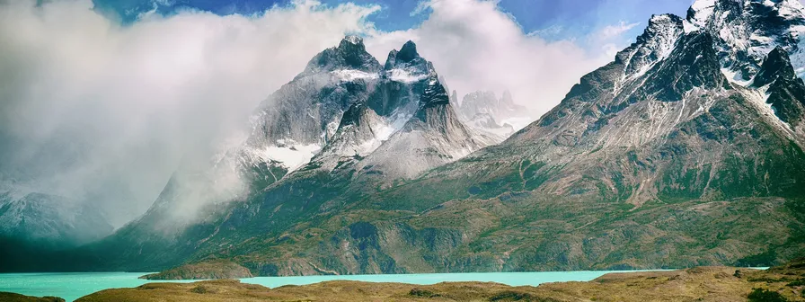 Jagged, snow-capped mountain peaks tower over a vibrant turquoise lake, with clouds partially obscuring the vast landscape. new Mapbox GL engine.